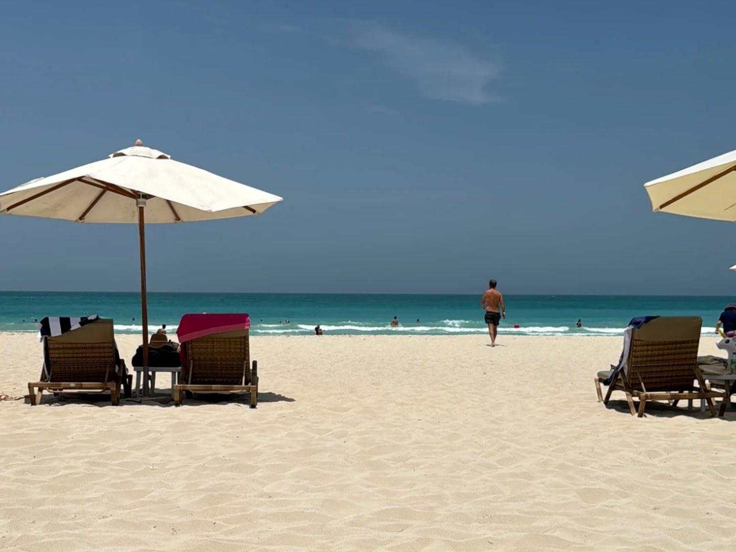 Cream parasol and two wooden sun loungers on white sand facing the turquoise sea in Abu Dhabi