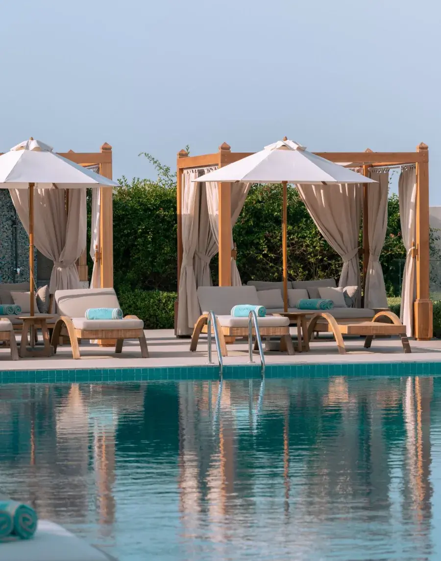 Curtained wooden cabanas with parasols and loungers beside a turquoise pool, in soft morning light
