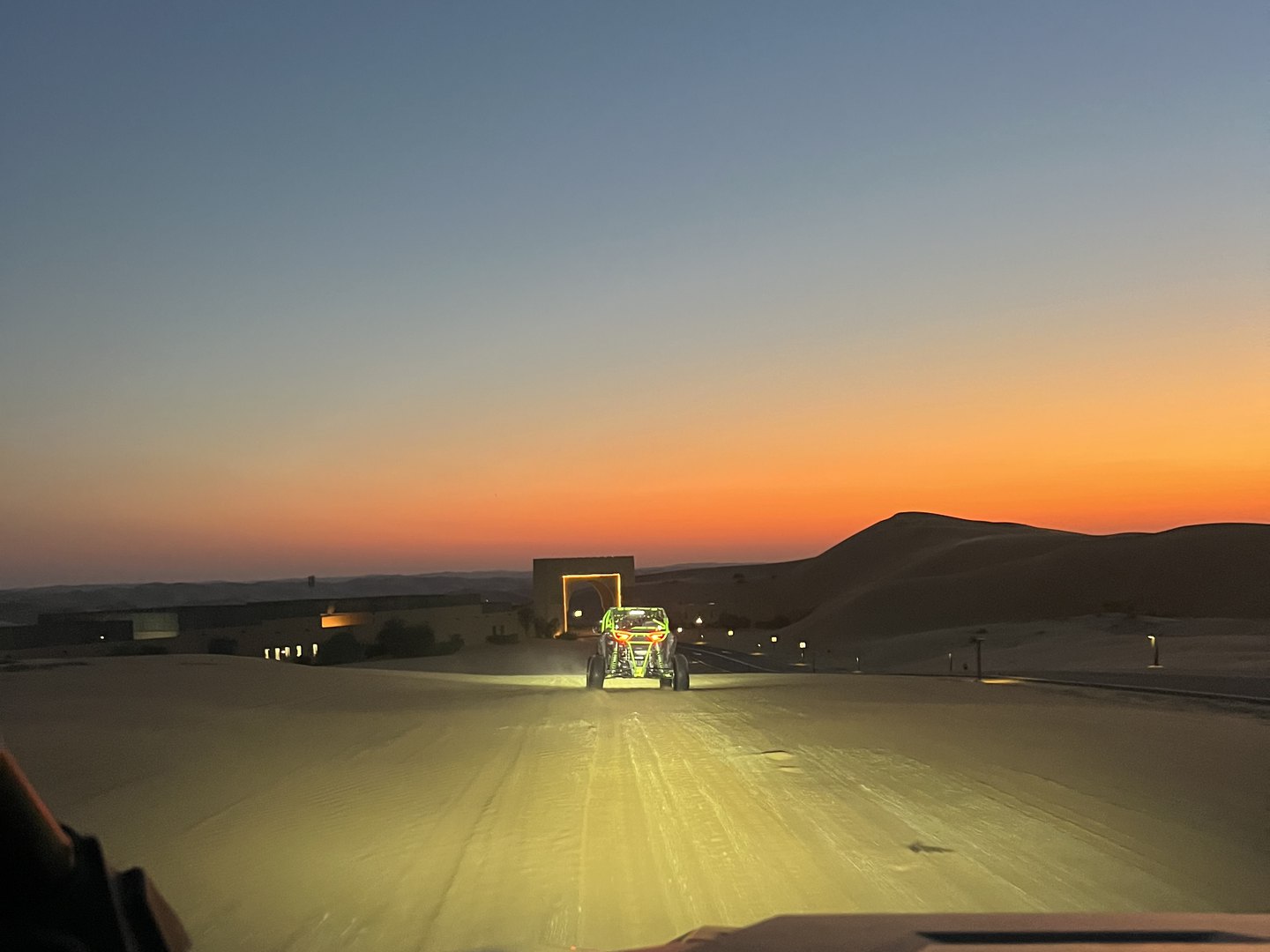 View from a dune buggy following another buggy across the sand at sunset, with the resort gate and dunes on the horizon