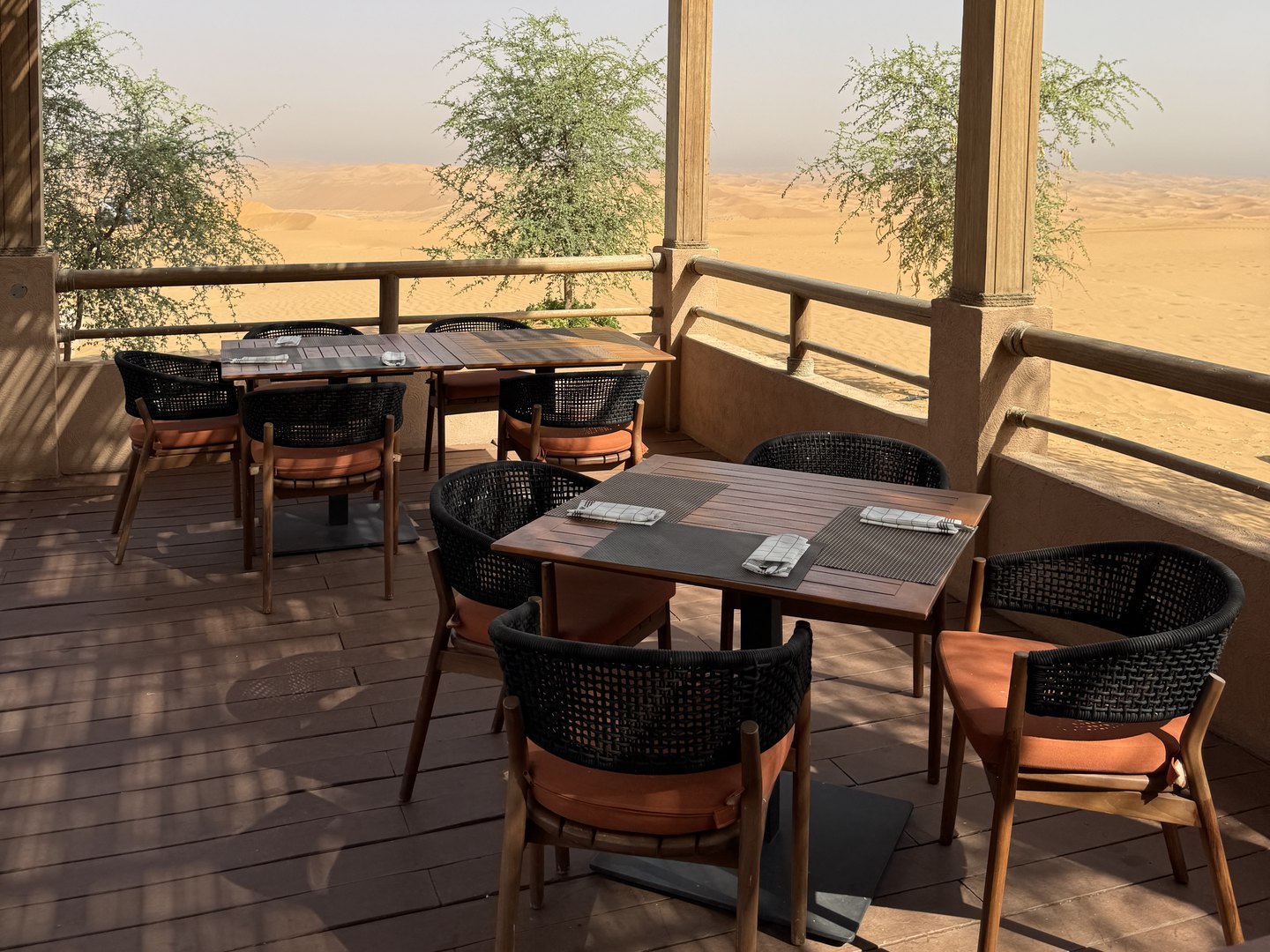 Shaded restaurant terrace with wooden tables and orange-cushioned chairs, overlooking the Liwa desert